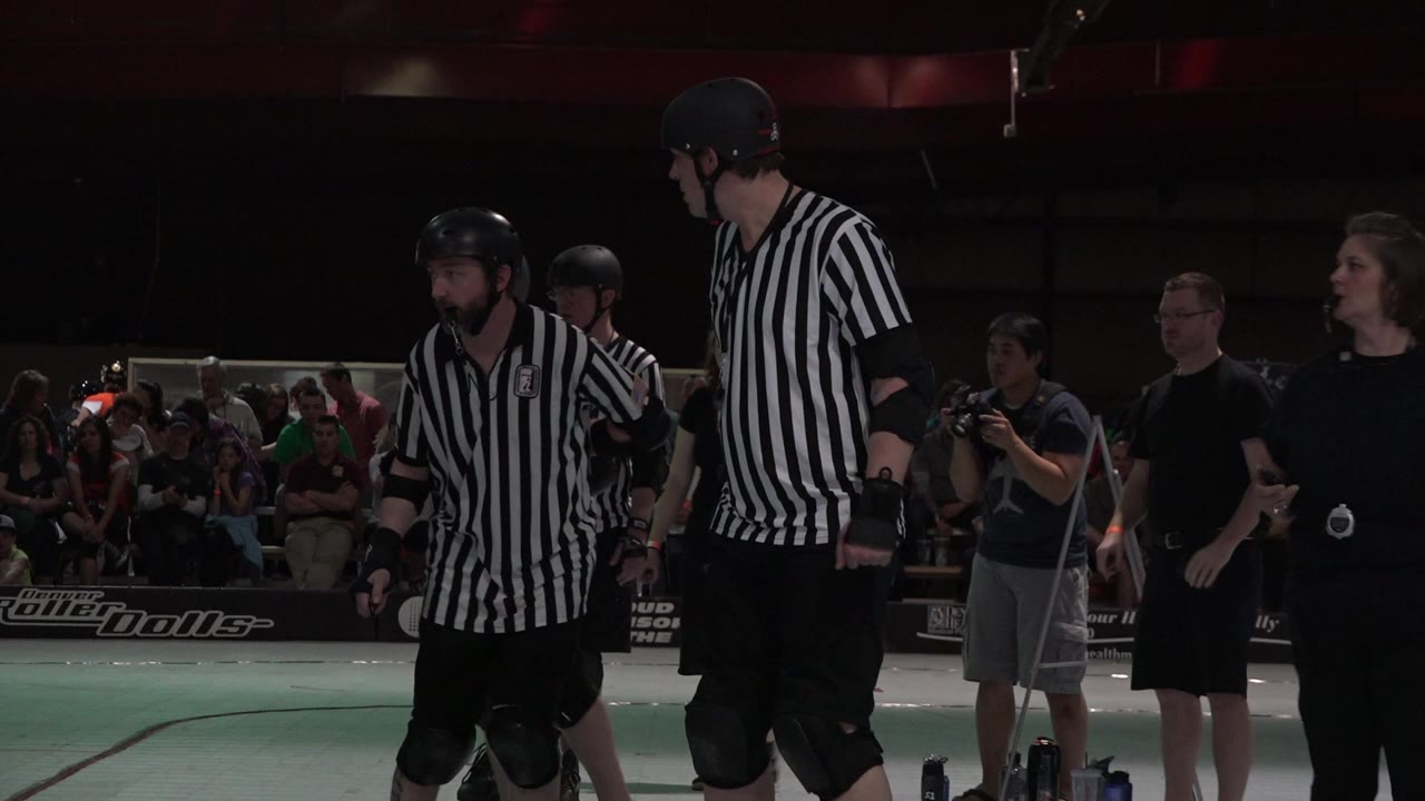 Roller derby referees stand near the track with spectators behind them.