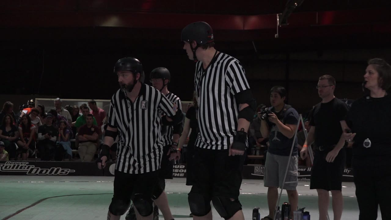 Two referees in striped shirts stand near the Denver roller derby track as spectators, a camera operator, and arena signage remain visible behind them.