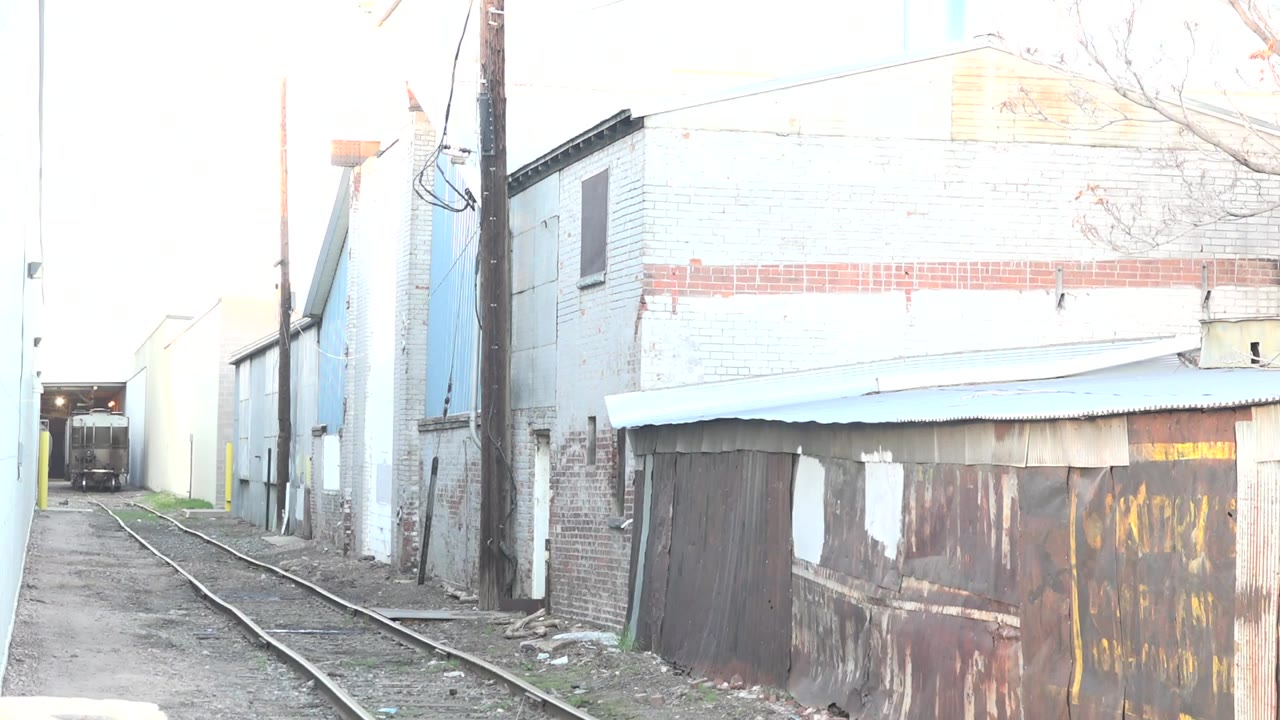 At 0:00 in Denver, rail tracks lead through a narrow alley bordered by pale brick, a utility pole, and a rusted shed wall, with no visible people.