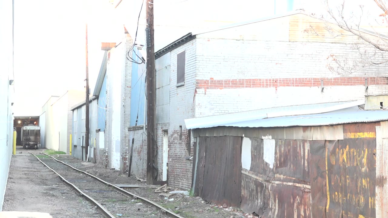 Ground-level Denver frame showing railroad tracks running past faded brick and metal-sided buildings in bright light, with no visible people.