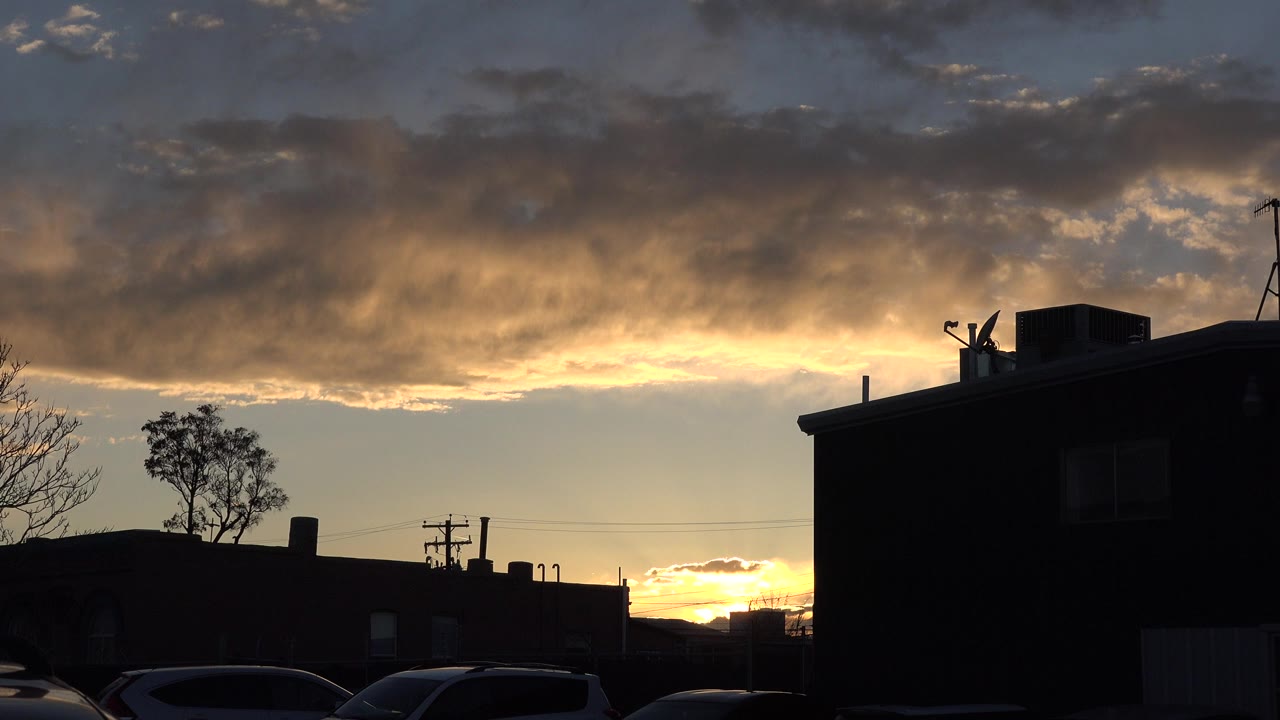 Sunset behind silhouetted rooftops and parked cars under clouds.
