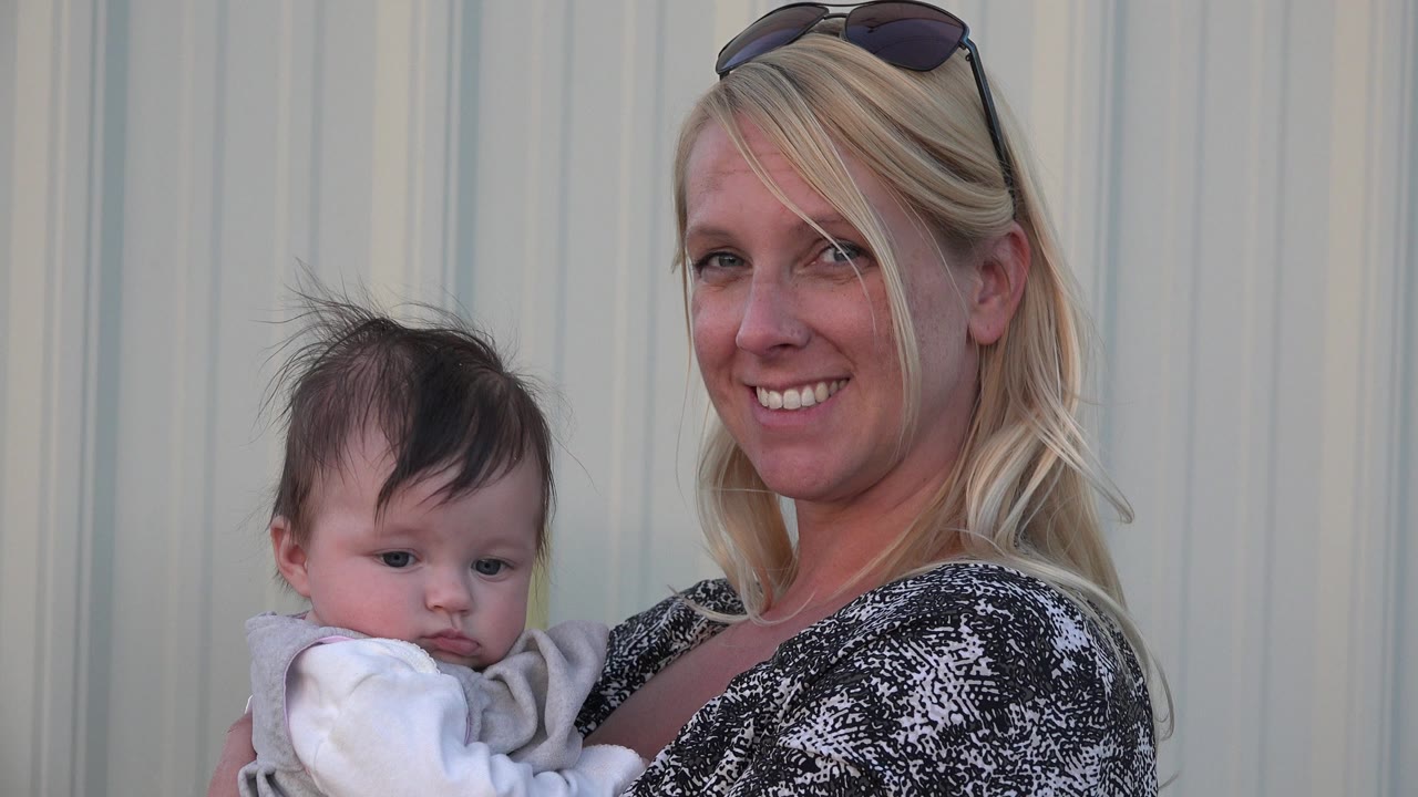 In Denver, Colorado, a blonde woman with sunglasses on her head holds a baby close while facing the camera against a pale vertical wall.