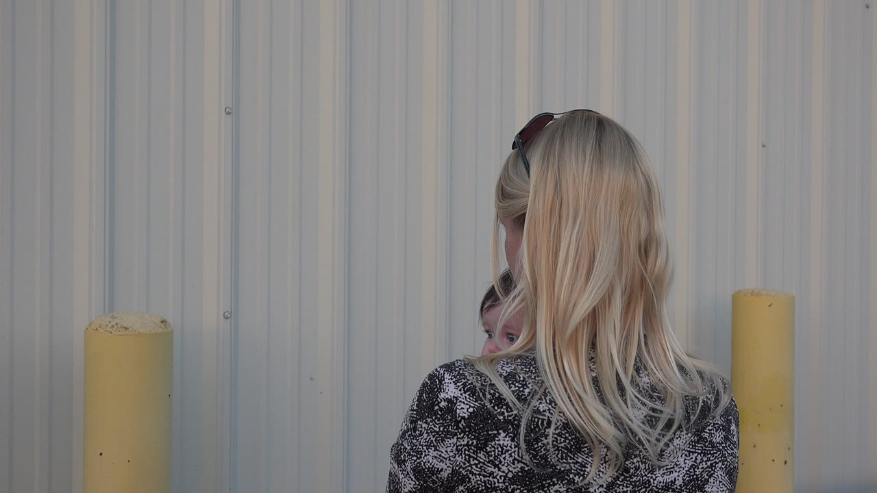 Blonde woman seen from behind holding a baby near a metal wall.