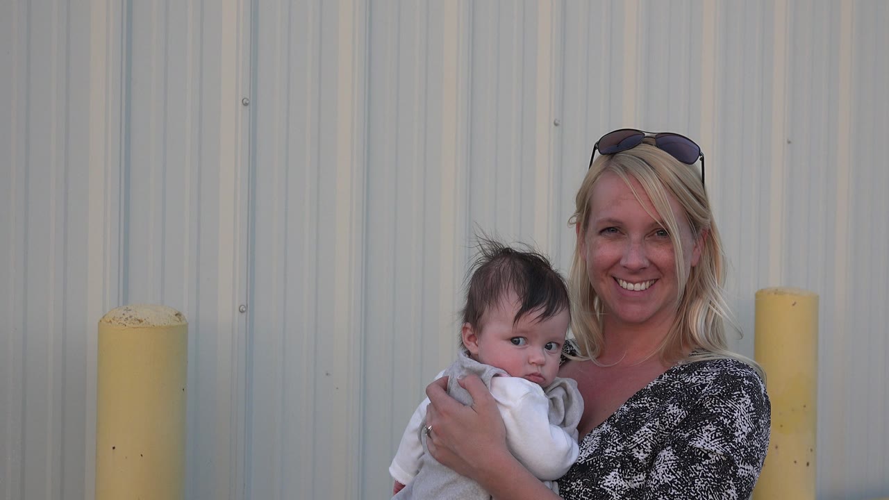 In Denver, Colorado, a blonde woman smiles while holding a baby close against a light corrugated metal wall with a yellow bollard at the side.