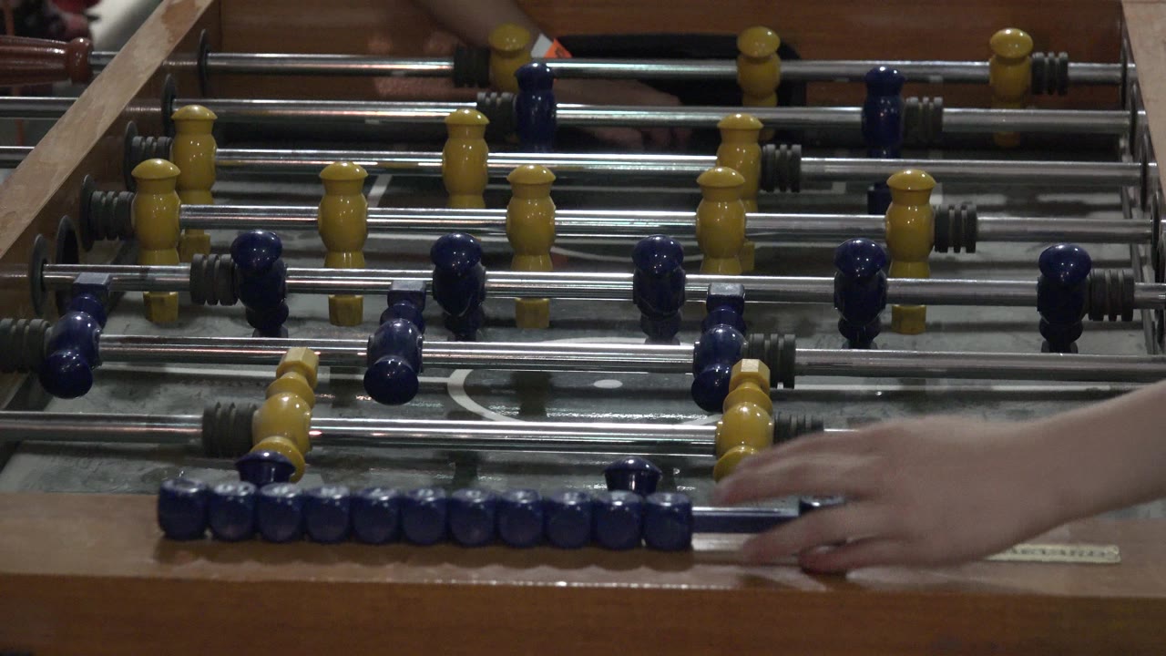 Denver 2015 frame with the hand crossing the front edge of the foosball table near clustered blue figures and yellow midfield pieces.