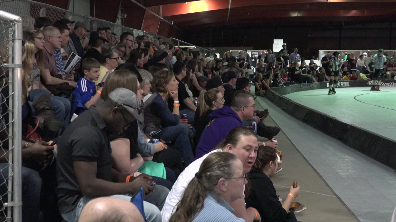 Crowd seated beside a roller skating ring with skaters in the distance.