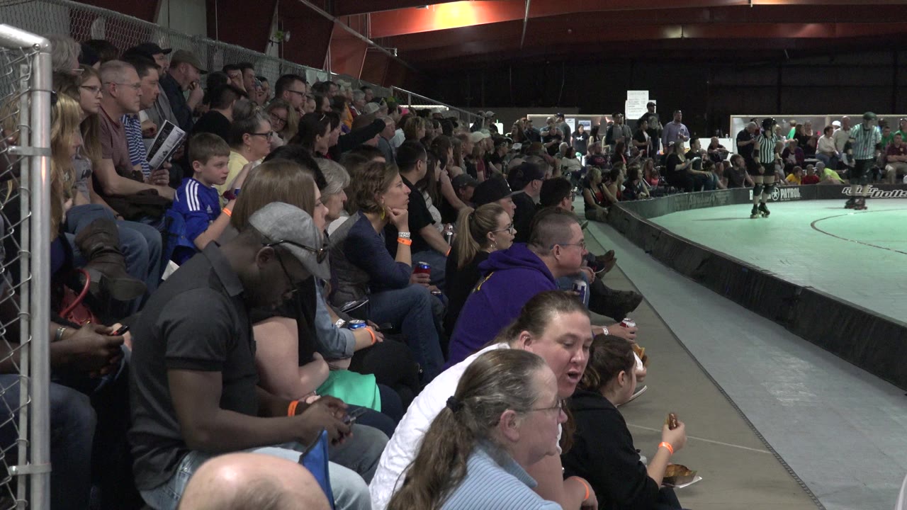 The Denver rinkside crowd remains seated along the fence, with a boy in a blue shirt near the aisle and skaters visible across the ring.