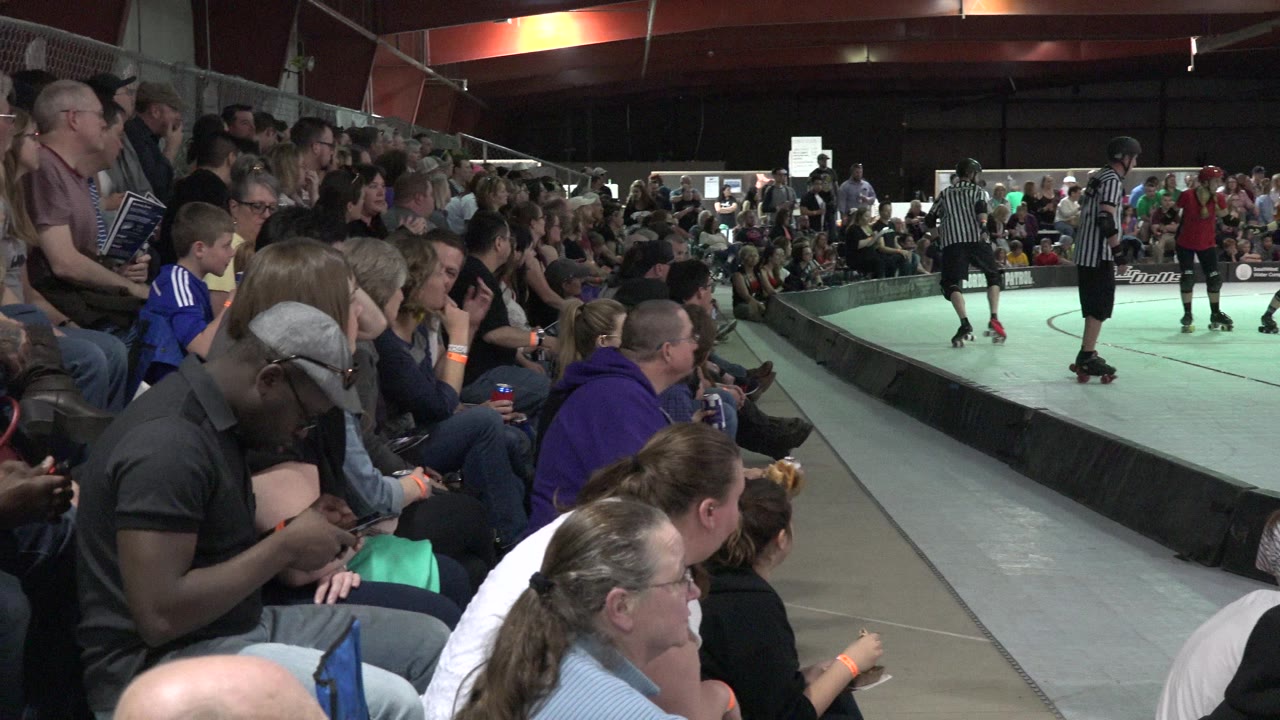 Spectators sit beside an indoor rink while roller skaters gather in the distance.