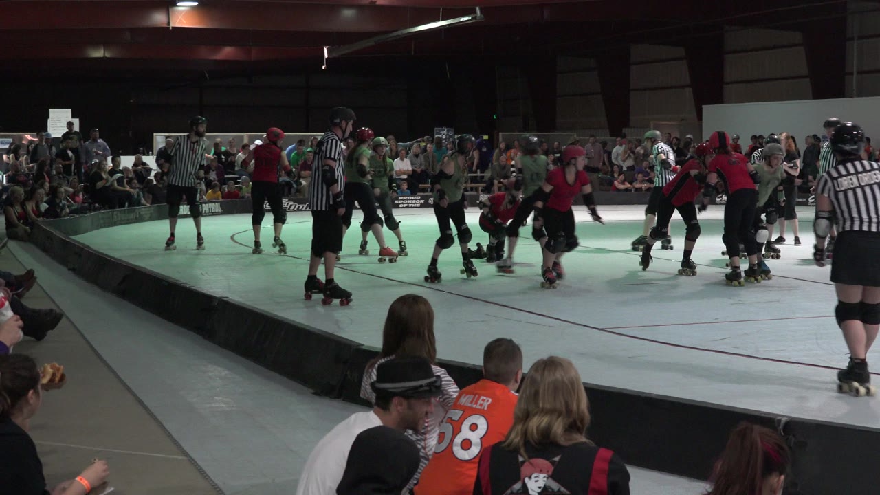 Roller derby skaters and referees on an indoor rink with spectators in the foreground.