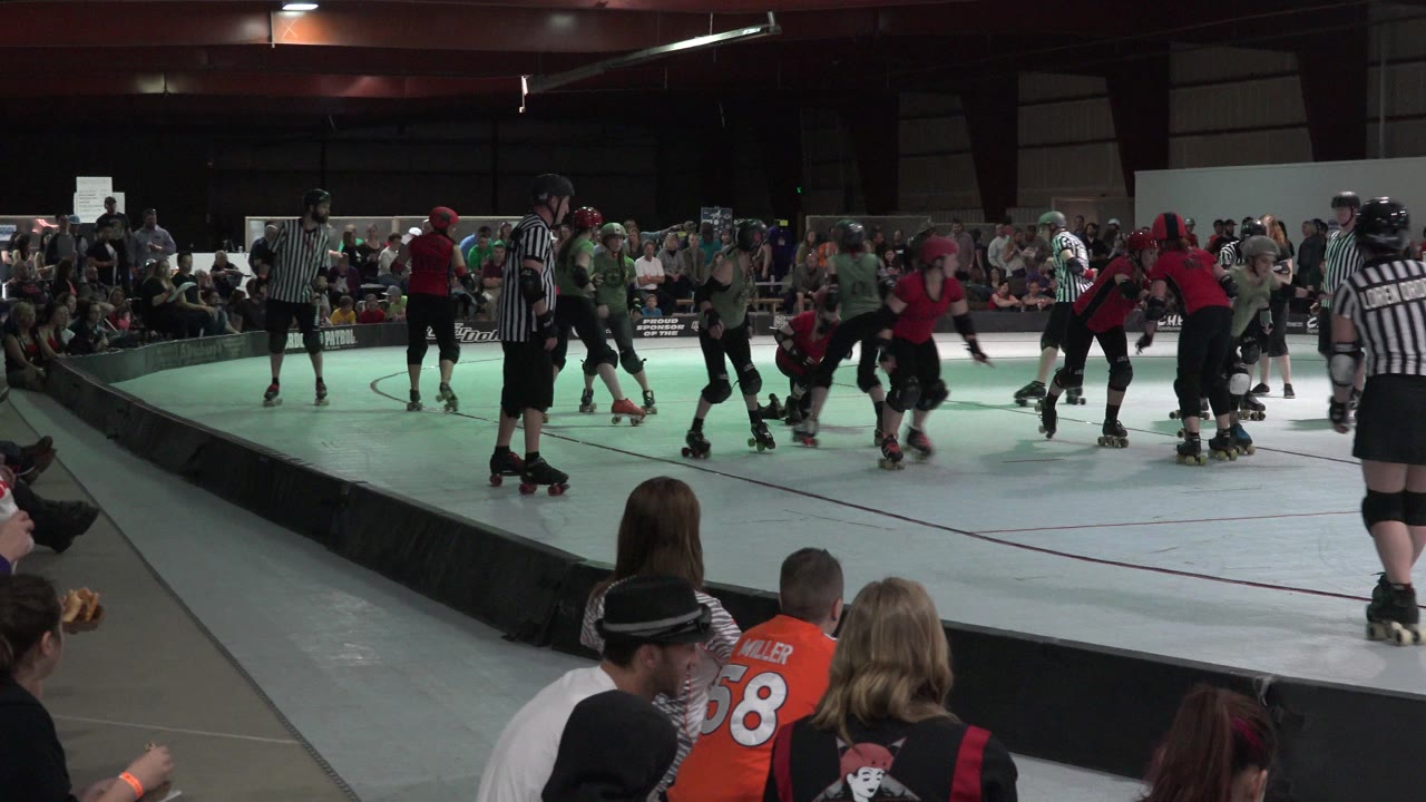 A rinkside view in Denver shows a referee near the camera, skaters forming up on the white track, and spectators watching from the far side and foreground.