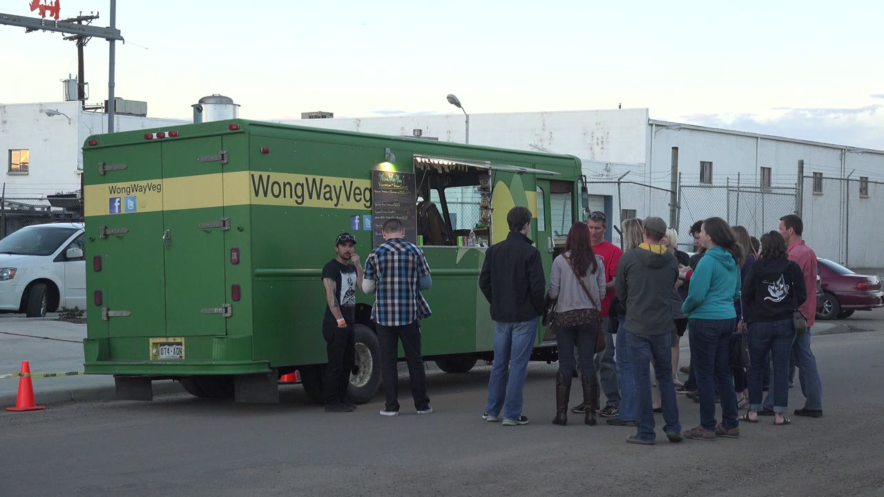 The green truck remains parked at the curb in Denver, Colorado, as several adults face the service window and others stand in a loose line on the street.