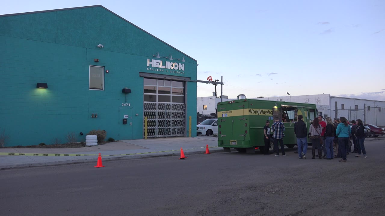 Green food truck outside a teal industrial building with people waiting nearby.