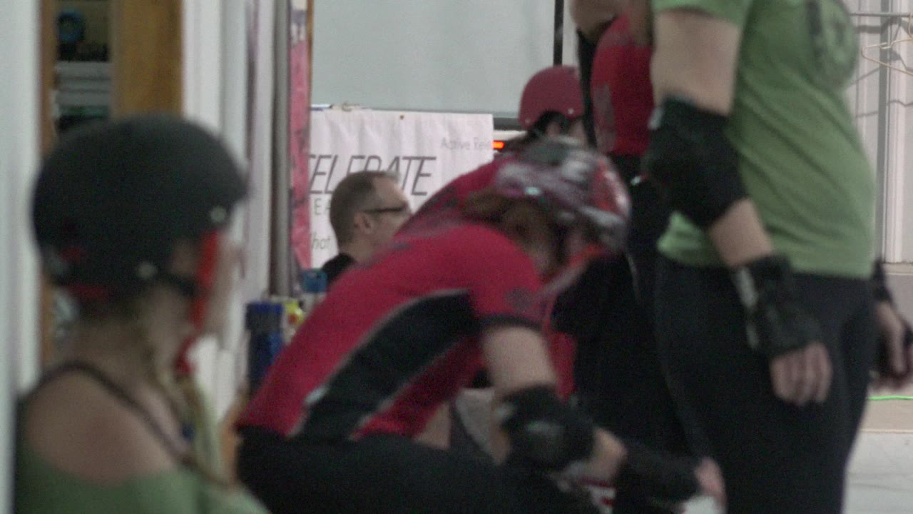 A red-and-black-uniformed skater bends forward near seated teammates in Denver, Colorado, with helmets and protective pads filling the bench-side frame.