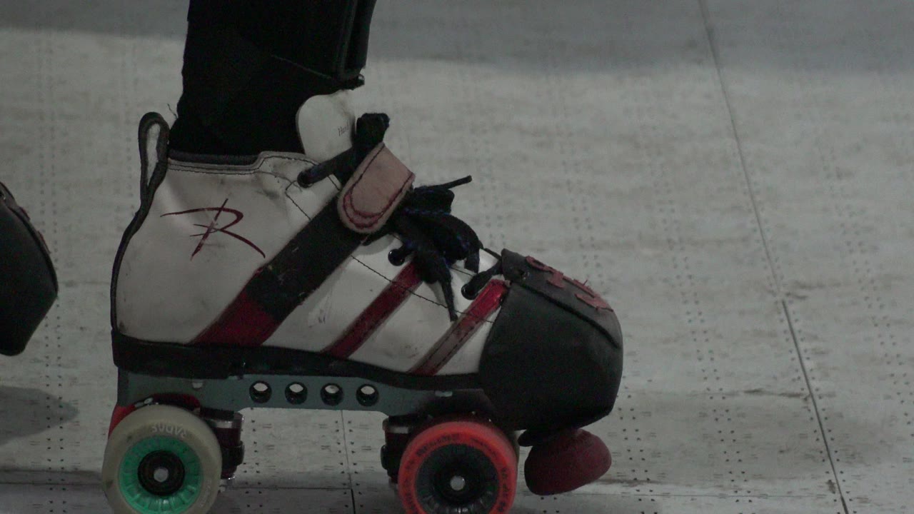 At 0:01, the same Denver skate remains in close side view, showing the black toe guard, red front wheel, green rear wheel, and marked pale floor, with no visible faces.