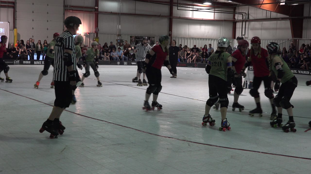 Denver roller derby match with a striped referee in the left foreground, skaters in red and light green jerseys clustered mid-rink, and spectators seated along the back wall.