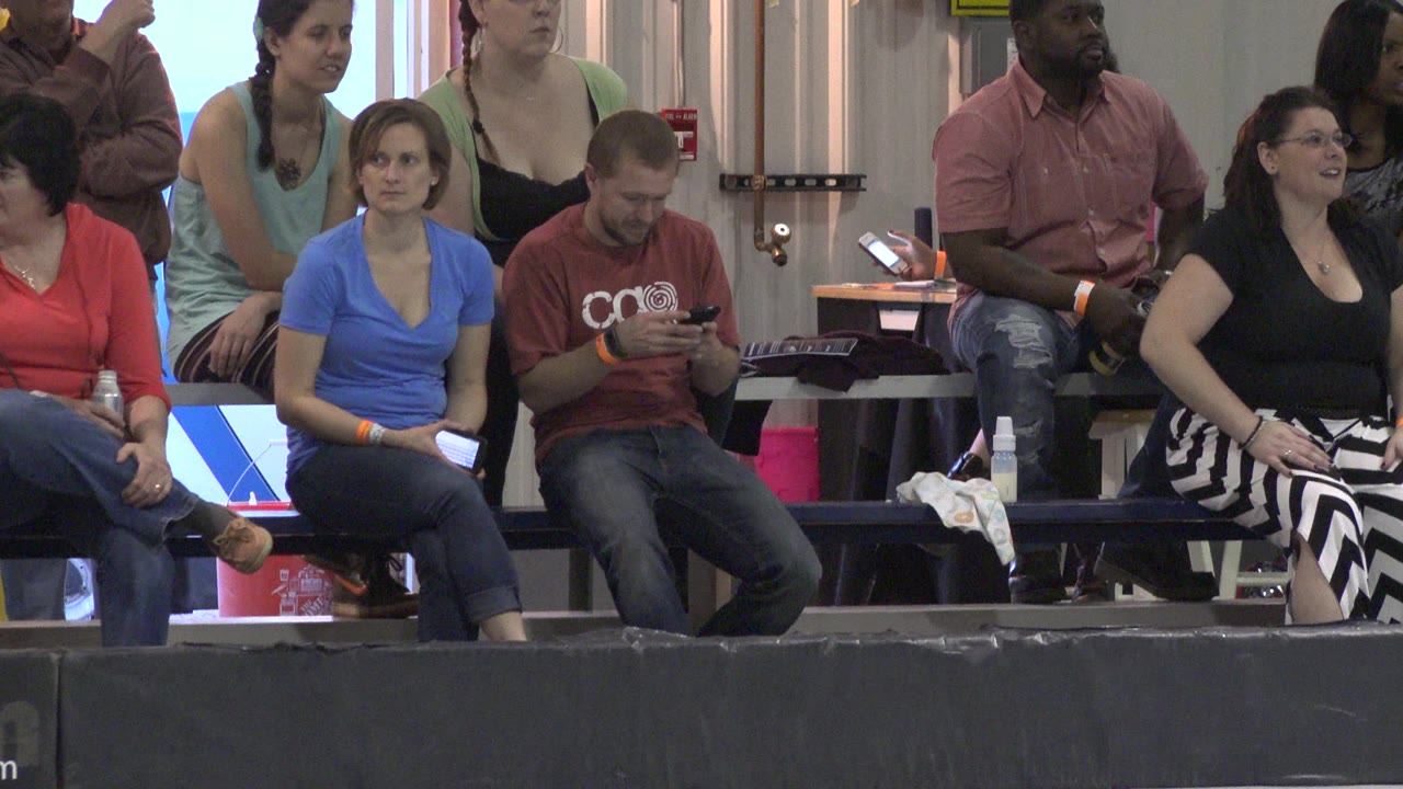 A Denver roller rink bleacher row shows seated adults, including a man looking down at a phone while others watch from behind the rink barrier.