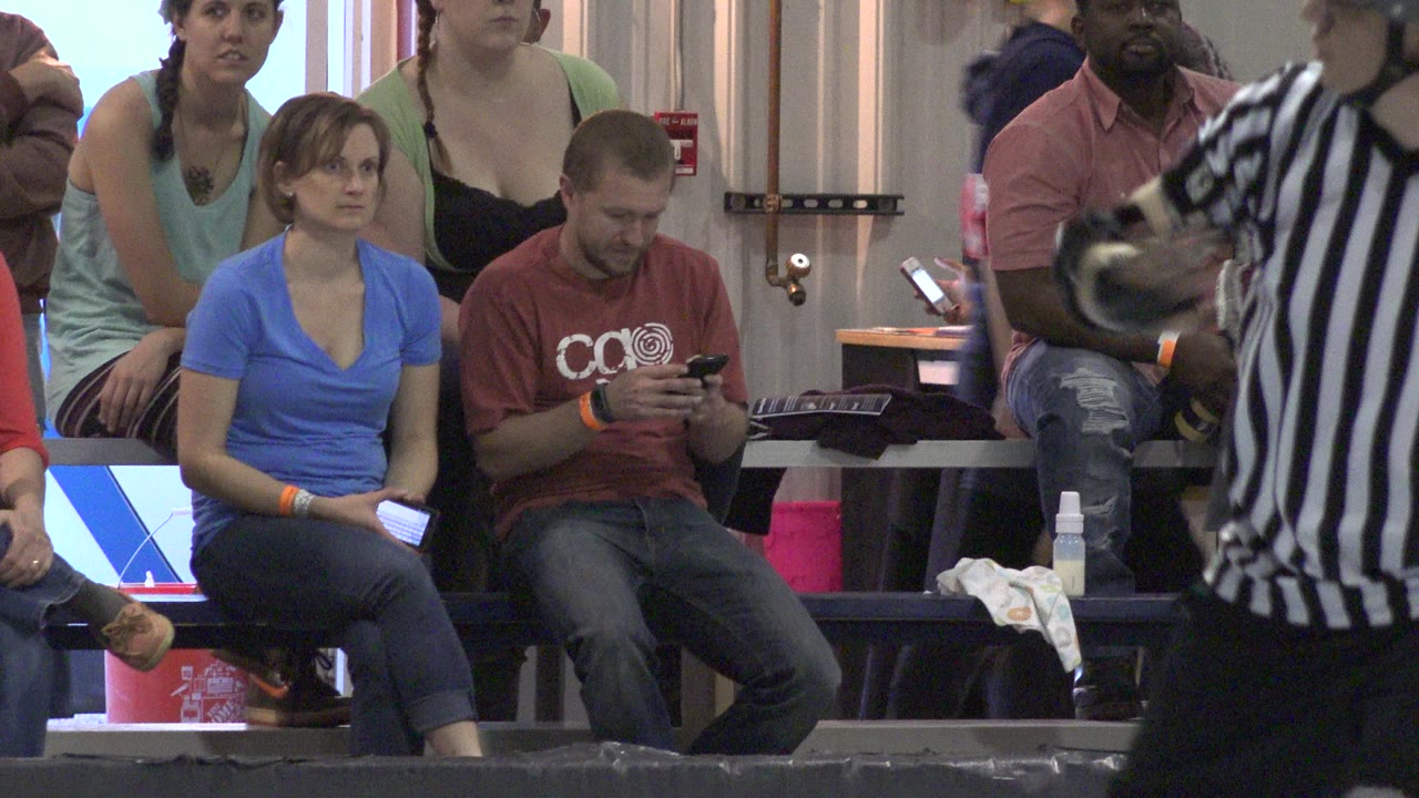 In Denver, Colorado, spectators sit on arena benches as a striped referee partially blocks the foreground and a man looks down at his phone.