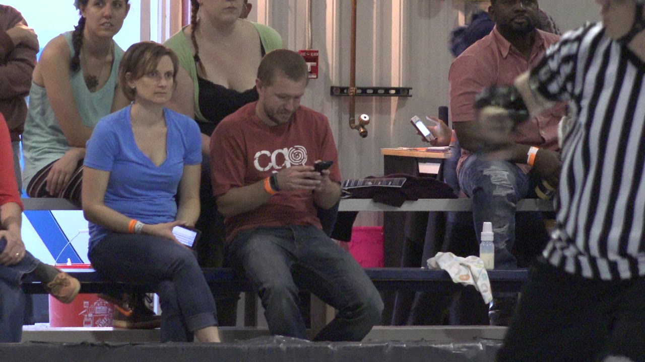 In Denver, Colorado, a seated man holds his phone beside a woman in blue while nearby spectators watch from the indoor roller derby seating area.