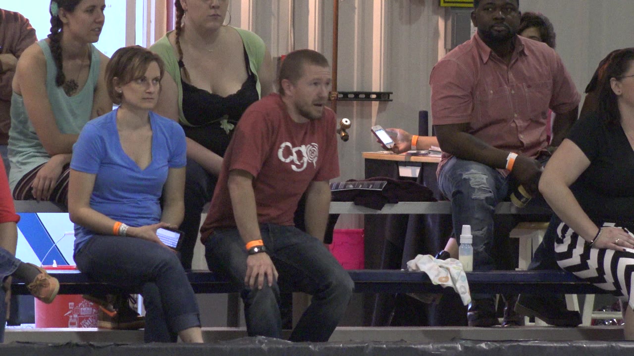 Adults sit on indoor bleachers in Denver, Colorado, with a woman in blue and a man in red facing forward while others watch from behind.
