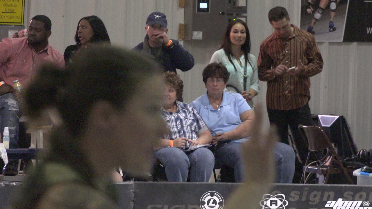 Spectators sit and stand behind an indoor event barrier while a blurred person crosses the foreground.