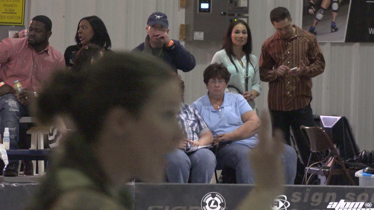 In Denver, Colorado, the indoor audience stays seated and standing behind the barrier while a blurred person moves across the foreground near center.