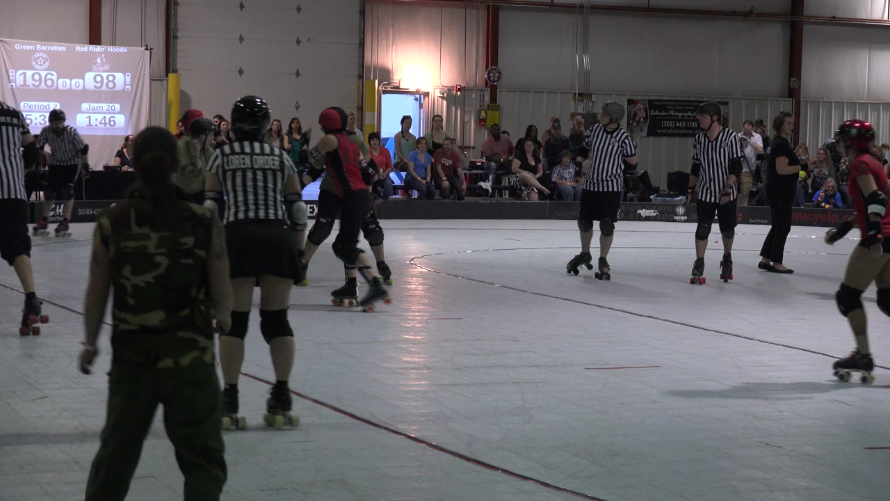 Denver indoor skating game with a striped referee and camo-clad skater in the foreground, players gathering mid-rink, and spectators seated behind a projected scoreboard.