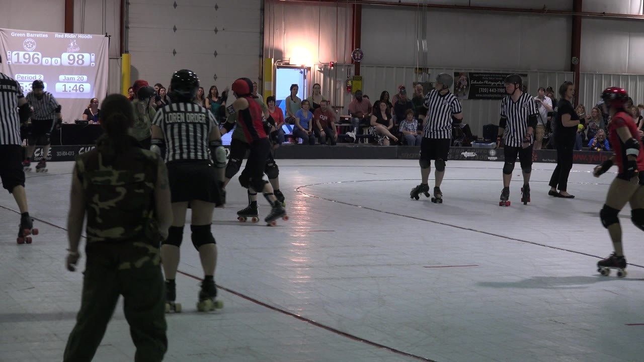 Denver rink scene with a black-helmeted referee near center, red and green uniformed skaters to the right, and spectators watching from benches along the far wall.