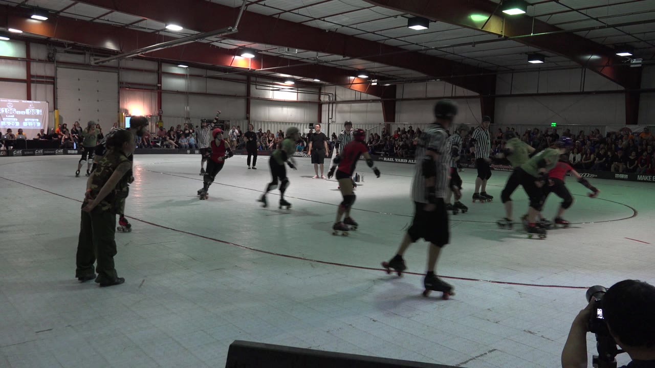 Roller derby skaters pass a referee in front of a seated crowd.