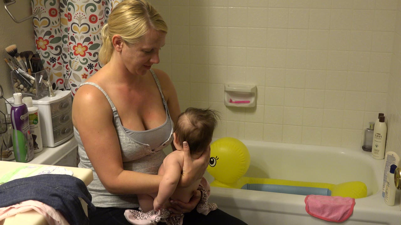 Woman holding infant beside bathtub with yellow inflatable duck tub.