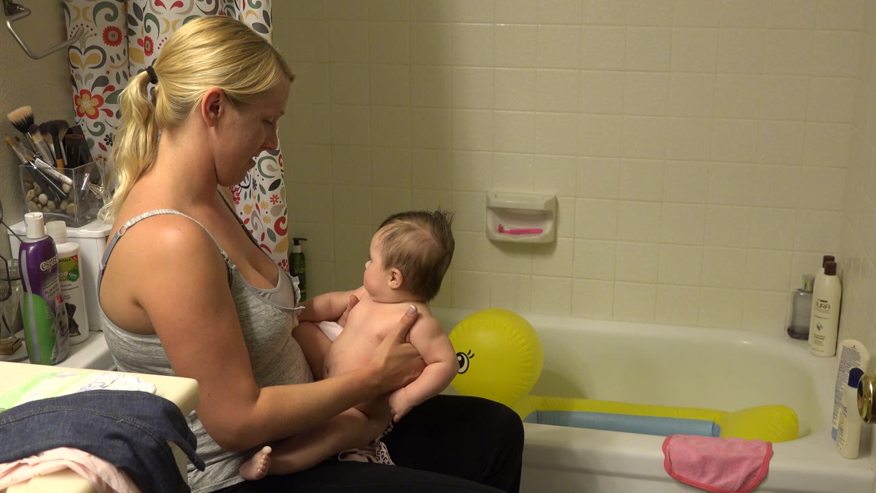 The mother holds the child facing the tub, with the yellow duck bath toy and bathroom bottles visible against the cream tile wall in Westminster, Colorado.