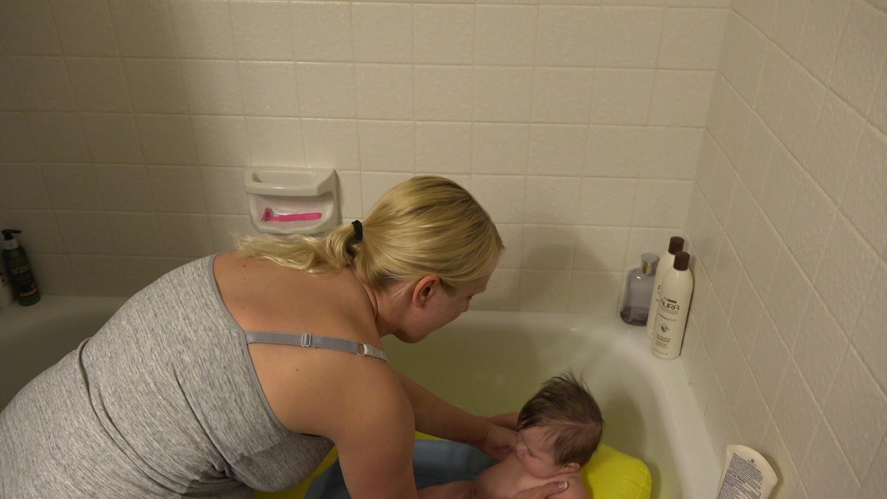 In Westminster, Colorado, a blonde mother leans over a bathtub toward an infant seated in a yellow and blue baby bath seat near cream tile walls.