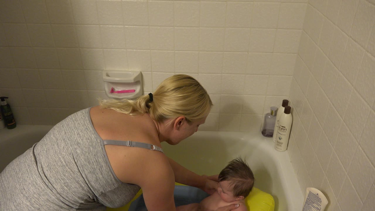 The mother bends closer over the white bathtub as the infant sits upright in the yellow bath support, with shampoo bottles visible along the tub edge.