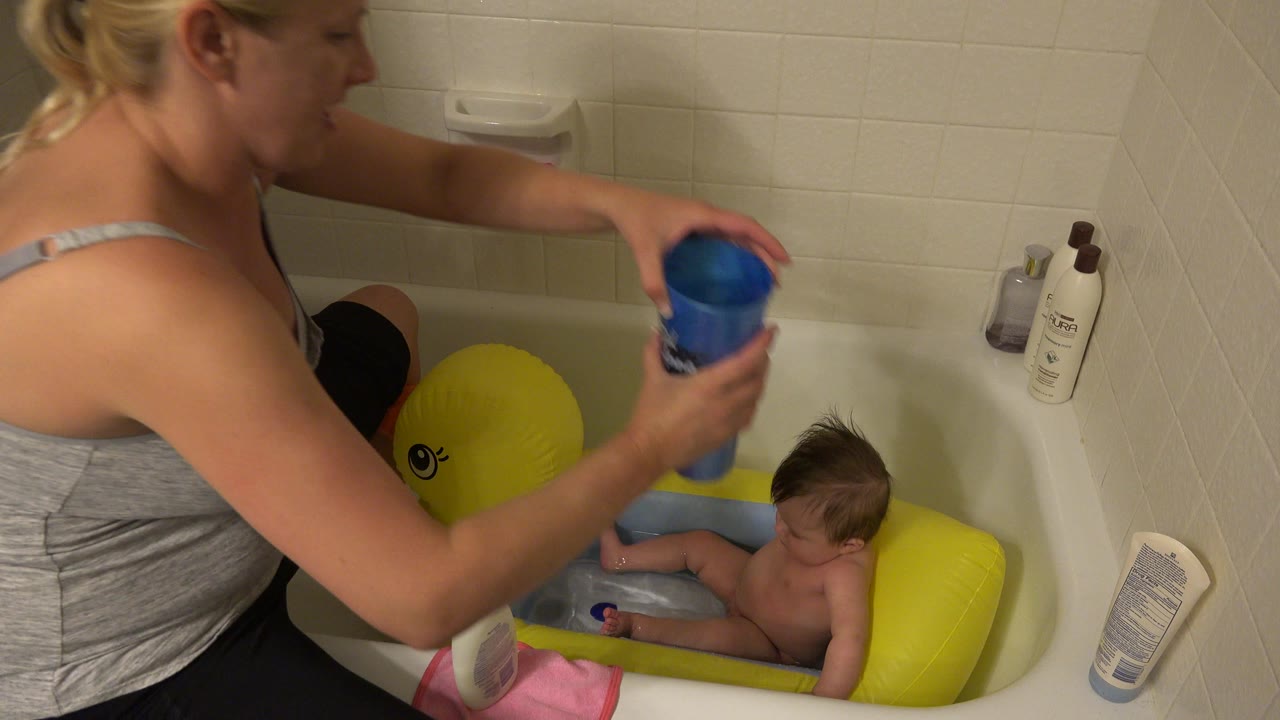 Blonde woman holds a blue cup over a baby in a bathtub.