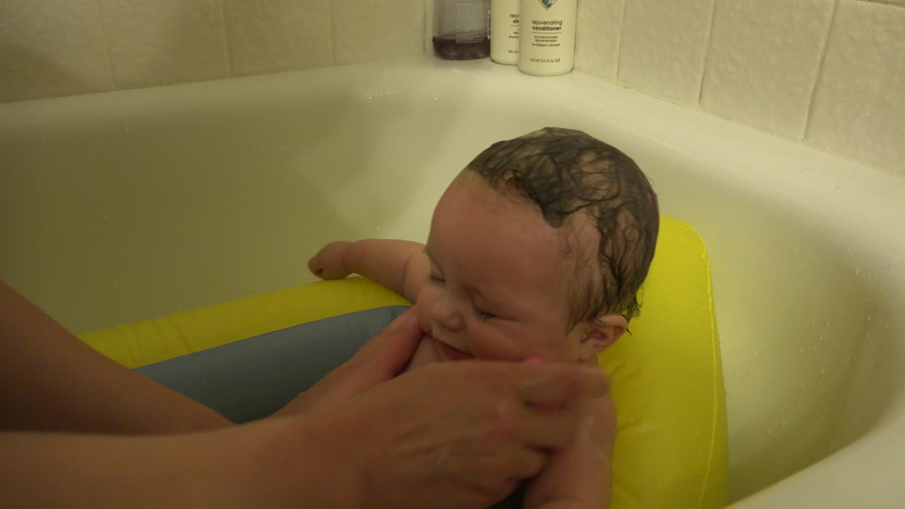 In Westminster, Colorado, the baby leans against the yellow side of the bath support as adult hands continue washing the baby's face and upper body.