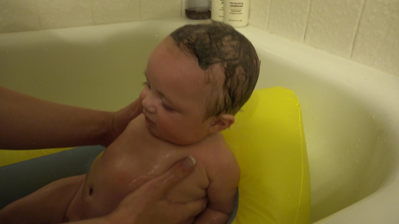 A wet infant sits in a bathtub in Westminster, Colorado, with an adult hand supporting the child's chest and a yellow bath seat behind them.