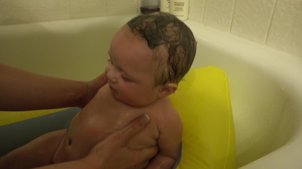 The infant looks downward in the Westminster bathroom tub while an adult hand holds the child's side near a yellow bath support.