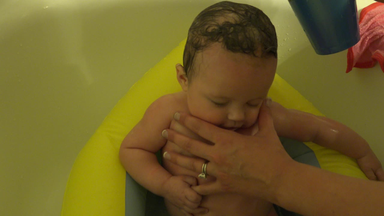 Infant in a yellow bath seat with an adult hand covering the mouth.