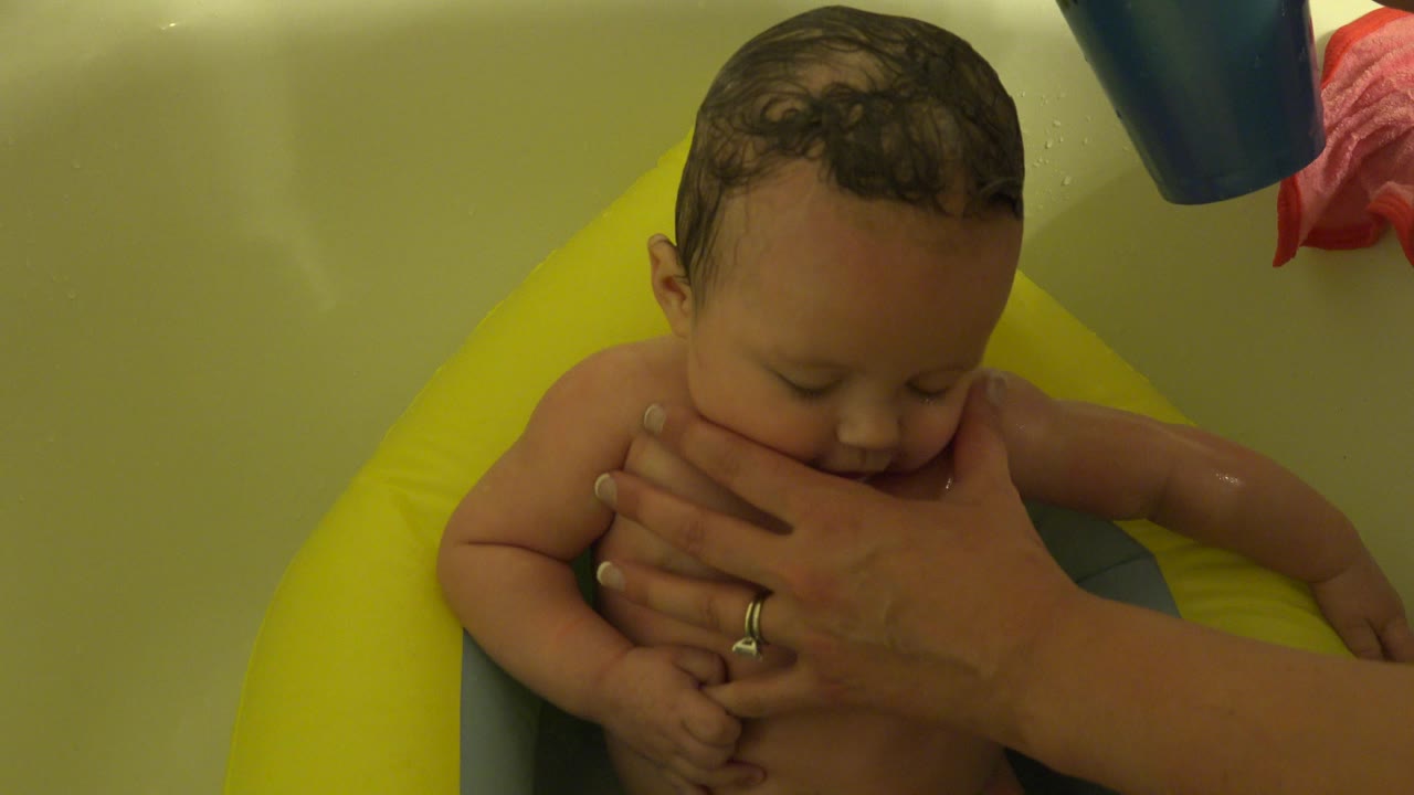 At 0:01 in Westminster, Colorado, the infant looks downward in the bath seat while the caregiver's hand rests across the lower face and chest.