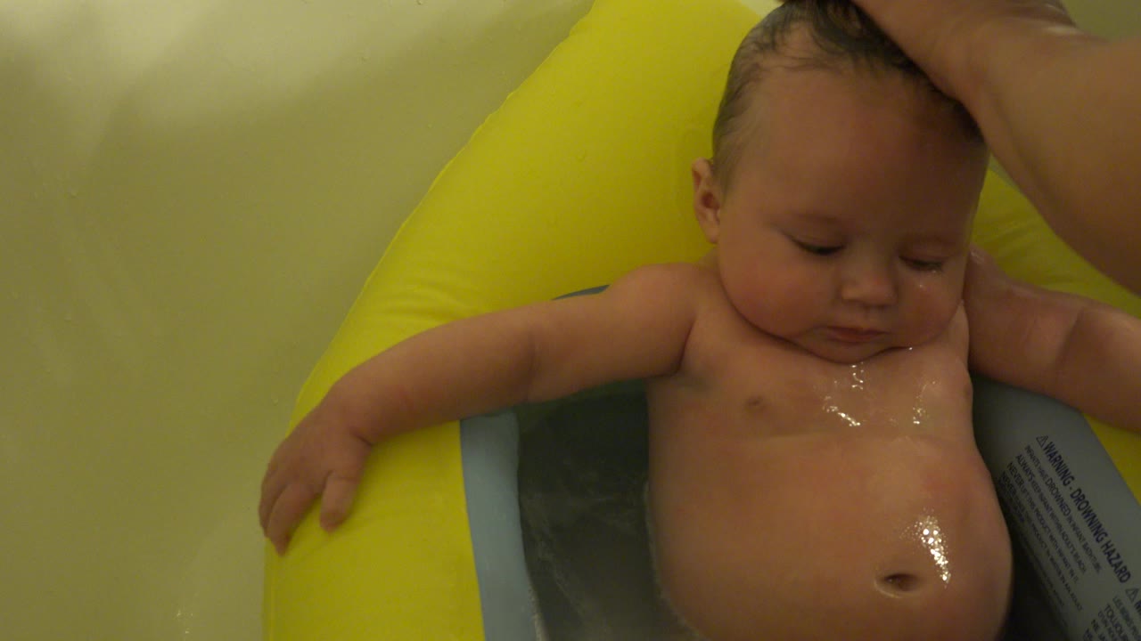 In Westminster, Colorado, a baby sits in a shallow bath on a yellow inflatable support while an adult hand rests on the child's wet hair at the start of the clip.