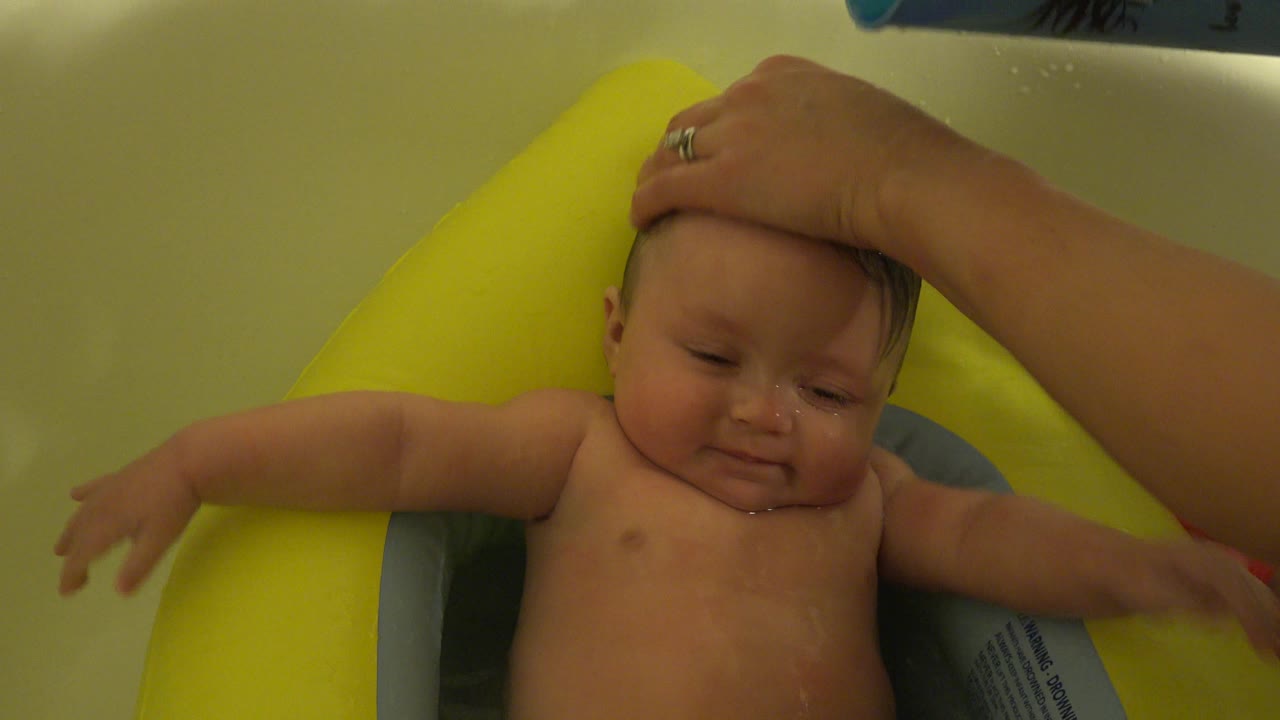 Wet baby in a yellow bath seat with an adult hand on the head.