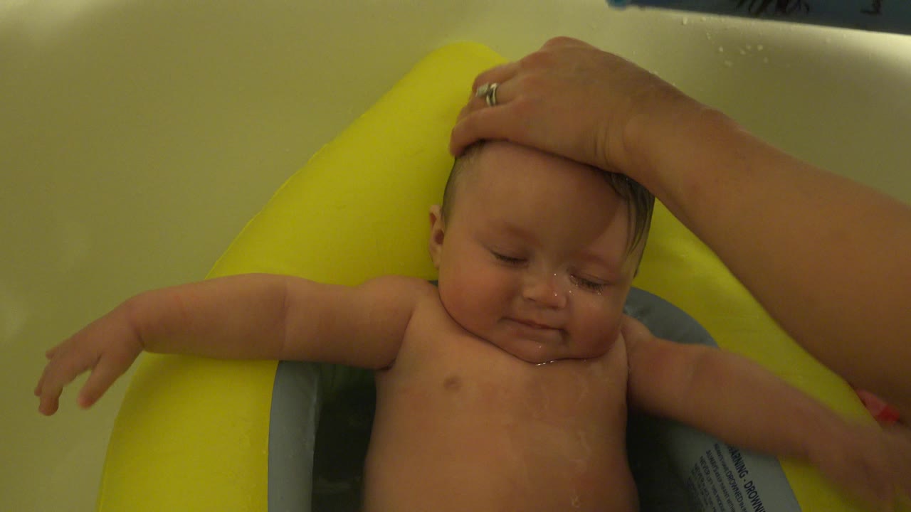 The baby remains in the yellow bath seat in Westminster, Colorado, with eyes closed and an adult hand placed across the top of the head.