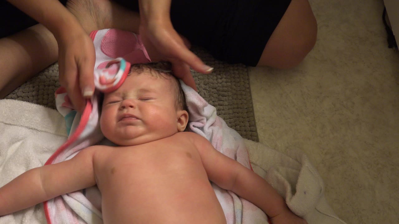 In Westminster, Colorado, the baby lies on a white towel while adult hands move the pink-trimmed towel across the baby's forehead and wet hair.