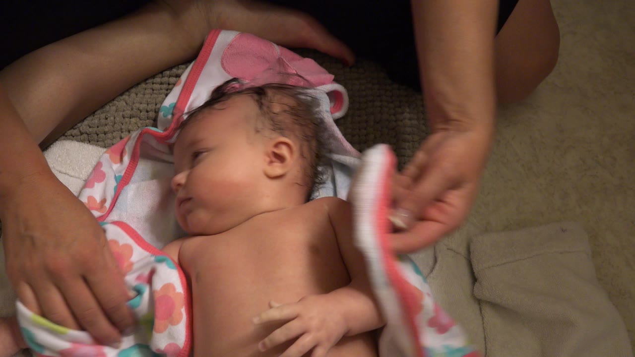 Baby lying on towels while adult hands hold a pink-trimmed towel.