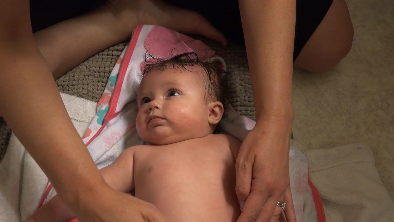 A baby lies on a pink-edged towel in a Westminster, Colorado home while an adult's arms reach around the child's torso at the start of the clip.