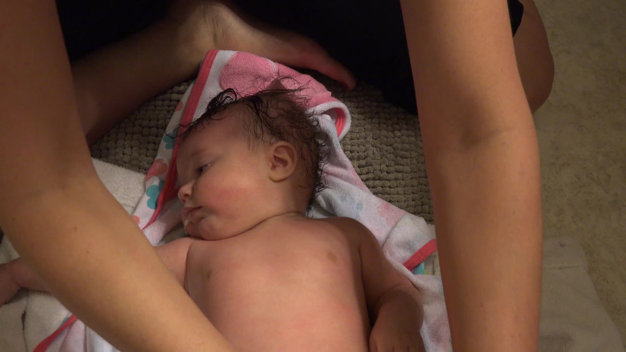 At 0:01, the baby remains on the towel with the adult's forearms close to the camera and the indoor floor visible around the mat.