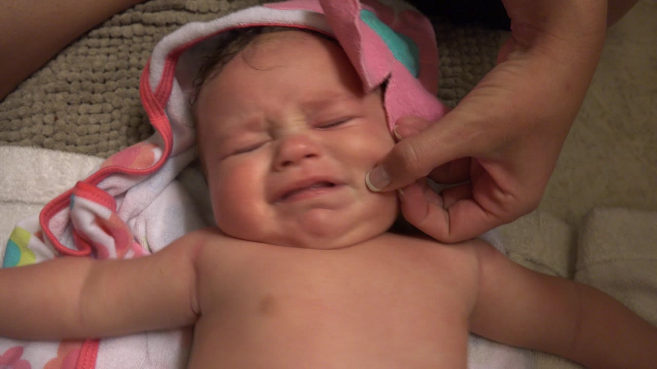 In Westminster, Colorado, the baby keeps eyes closed while crying, with a parent's fingers pressed gently near the cheek and towel edge.