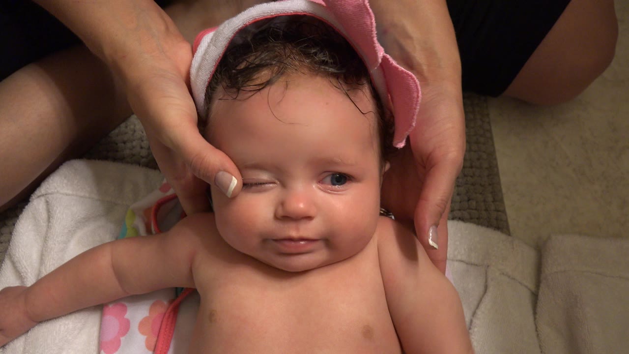 In Westminster, Colorado, a baby lies on a bathroom floor towel while adult hands steady a pink hooded bath towel around the baby's damp hair.