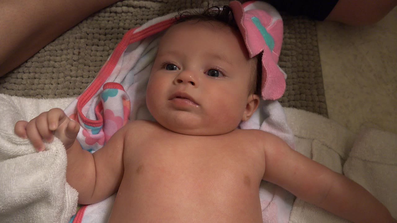 At 0:01, the baby remains centered on the towel in Westminster, Colorado, with one hand raised slightly and the pink bath blanket tucked near the head.