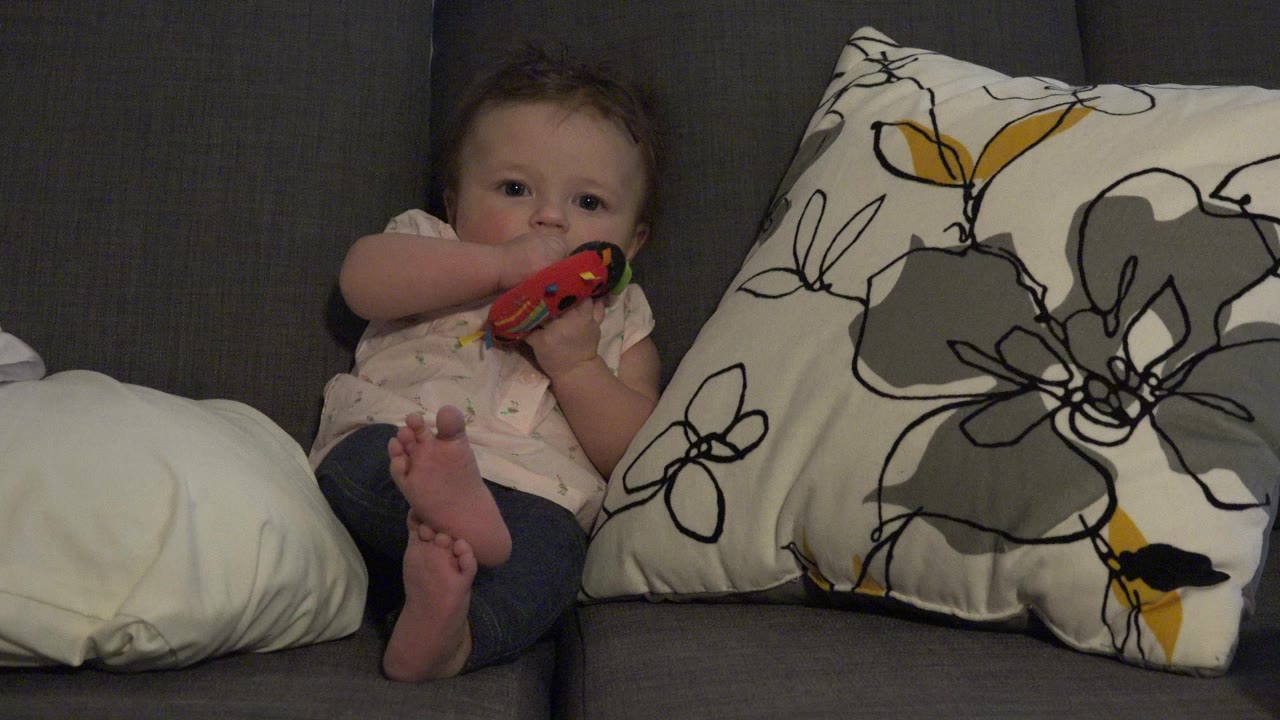 Baby sitting on a gray sofa holding a red object near the mouth.