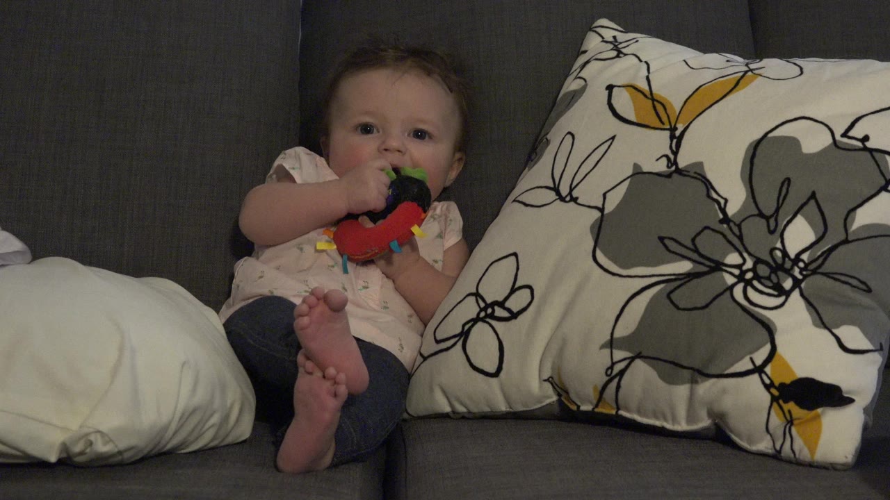 At 0:00 in Westminster, Colorado, a baby sits on a gray couch with bare feet crossed, holding a colorful toy or bottle-like object up to the mouth beside patterned pillows.